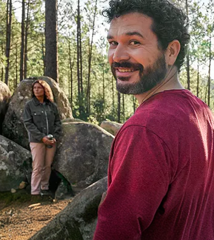 Representación actuada de un hombre adulto de al menos 19 años de edad sonriendo en un bosque y unamujer sentada en unas rocas en el fondo