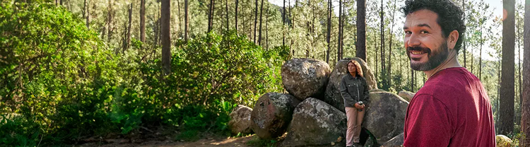 Representación actuada de un hombre adulto de al menos 19 años de edad sonriendo en un bosque y unamujer sentada en unas rocas en el fondo
